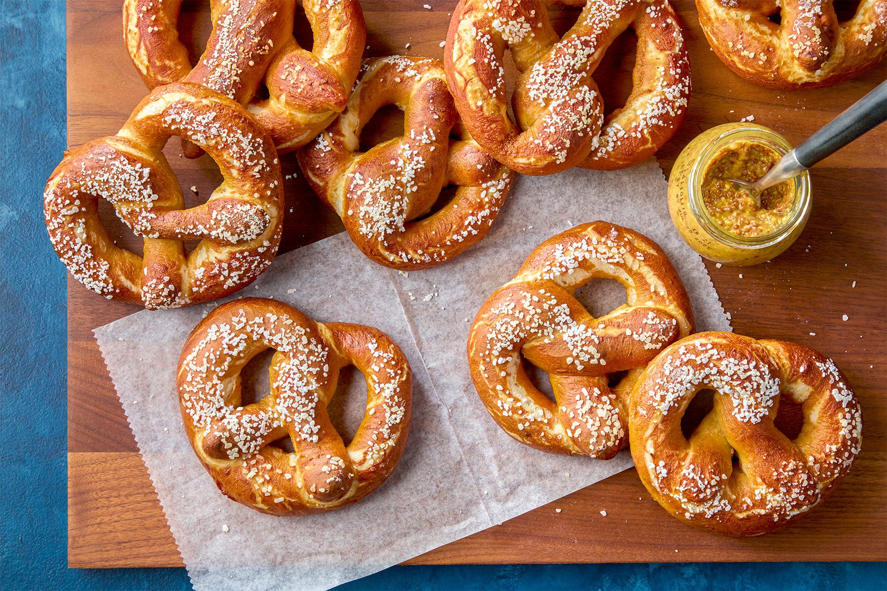 A wooden board displaying freshly baked pretzels sprinkled with coarse salt. Two pretzels rest on parchment paper, and a small pot of mustard with a spoon sits to the side. The background is a deep blue.