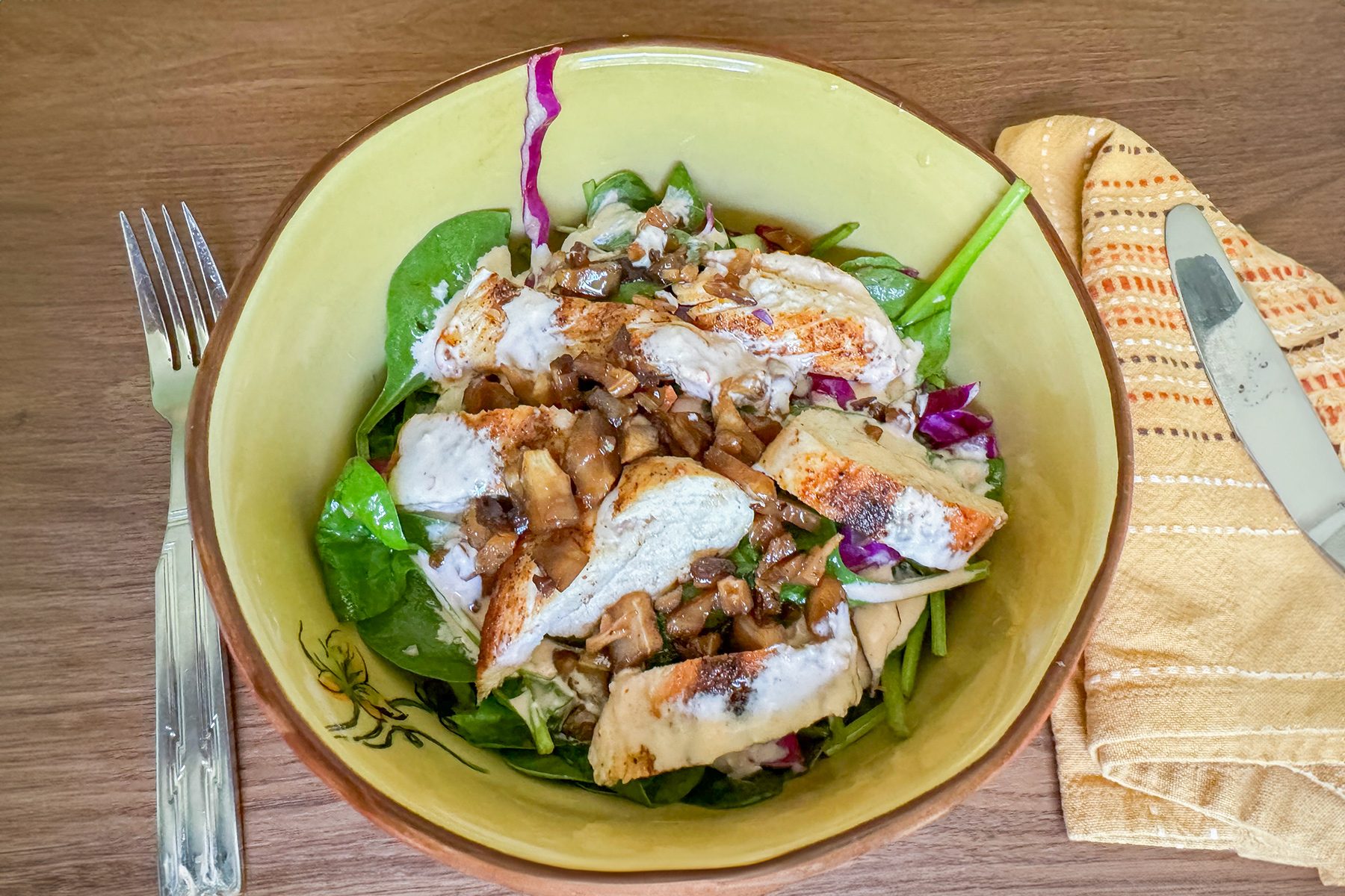 A bowl of salad with sliced grilled chicken, lettuce, and purple cabbage, topped with creamy dressing. A fork and knife are placed beside the bowl on a wooden table, along with a folded yellow napkin.
