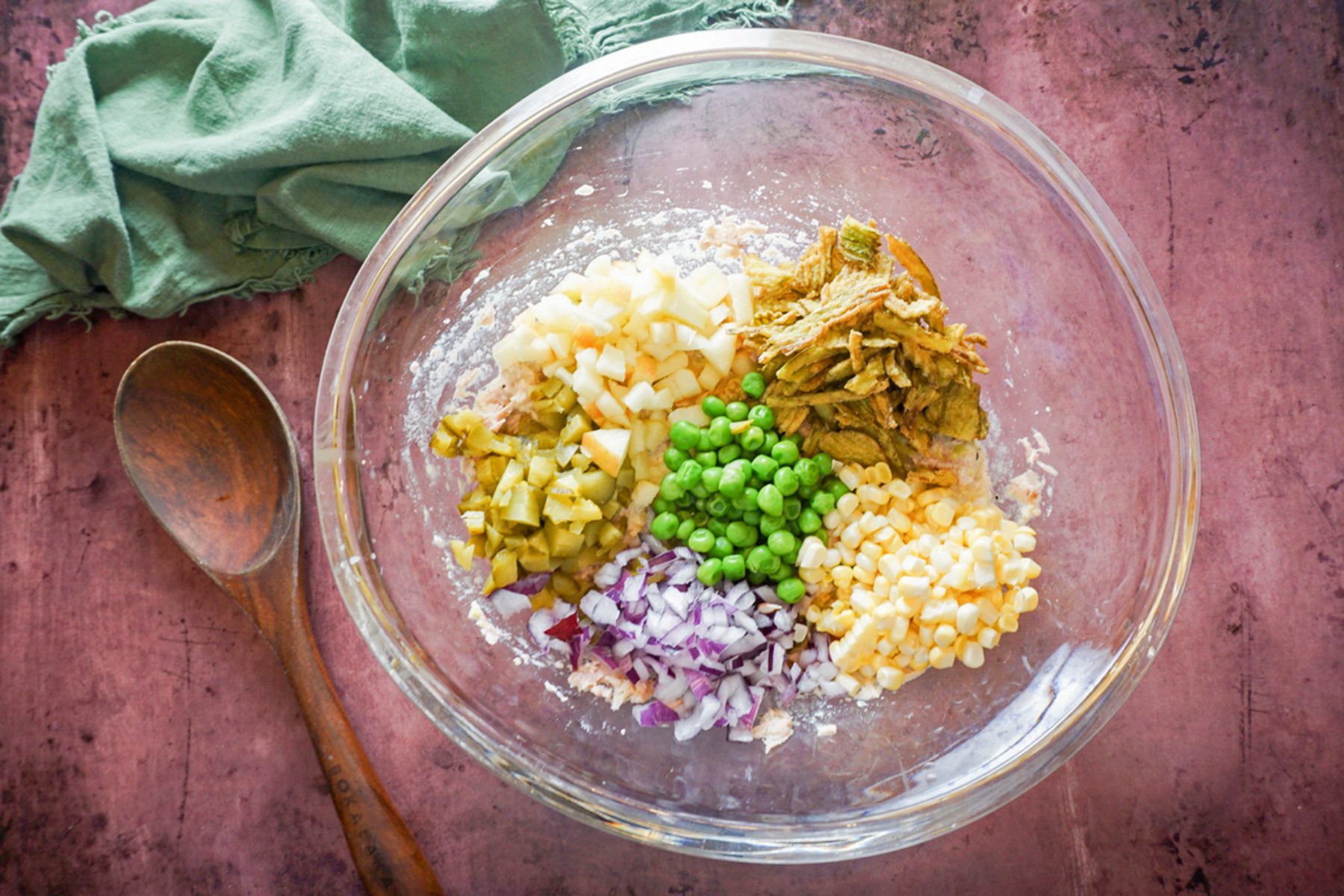 A clear glass bowl on a pink surface contains chopped ingredients: onions, canned tuna, green peas, corn, and red onions. A wooden spoon lies beside the bowl, with a green cloth in the background.