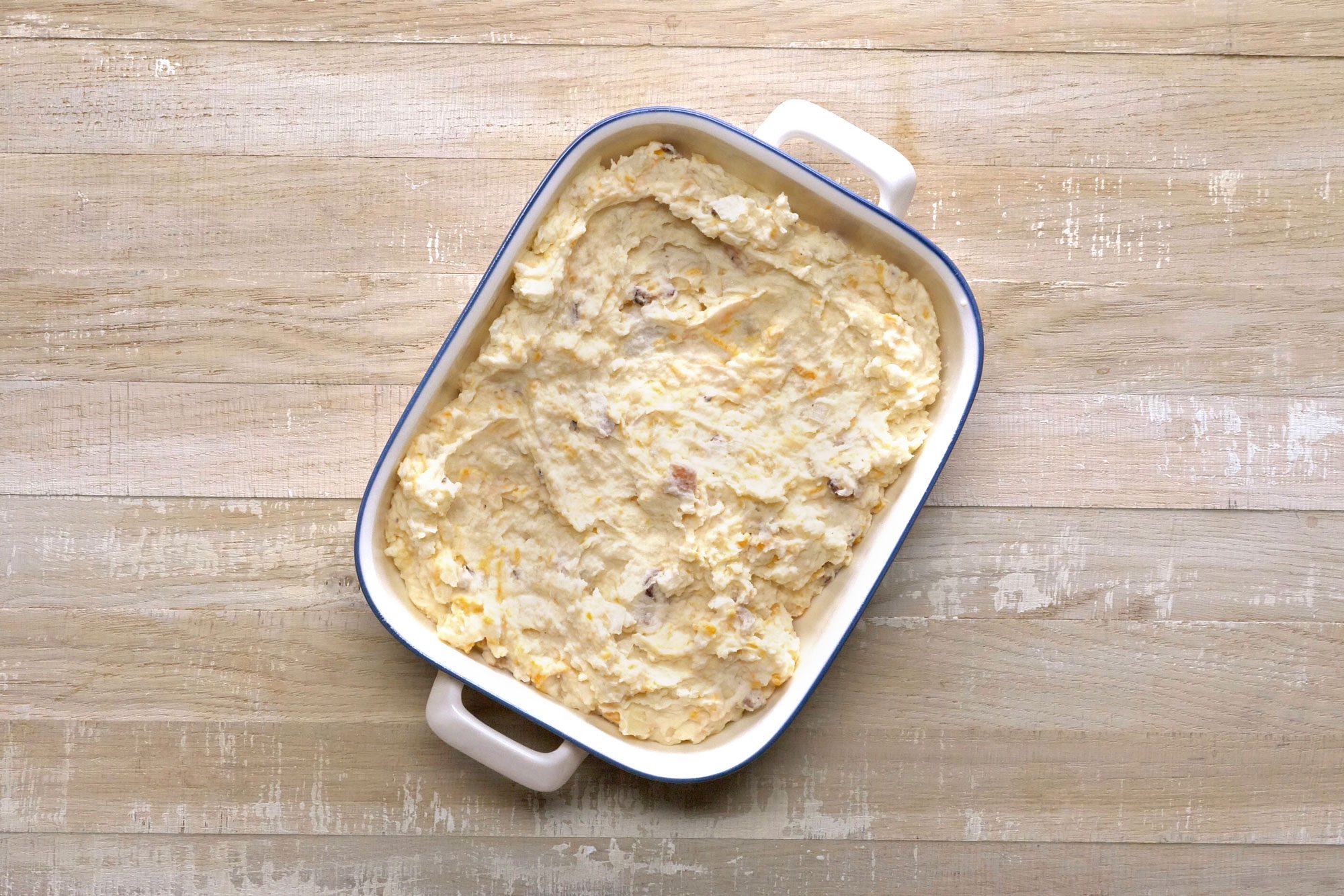 A baking dish filled with creamy, yellow mashed potatoes is set on a light wooden table. The surface is smooth with some texture visible, indicating it has been recently prepared. The dish has handles on each side.