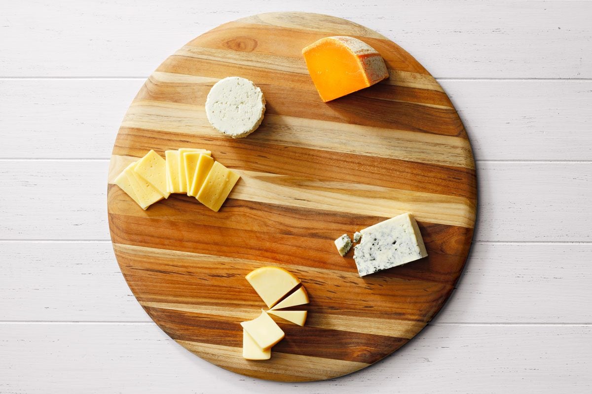 A round wooden board displaying a variety of sliced and whole cheeses, including a wedge of orange cheddar, a round of soft white cheese, and slices of yellow cheese, on a white wooden surface.