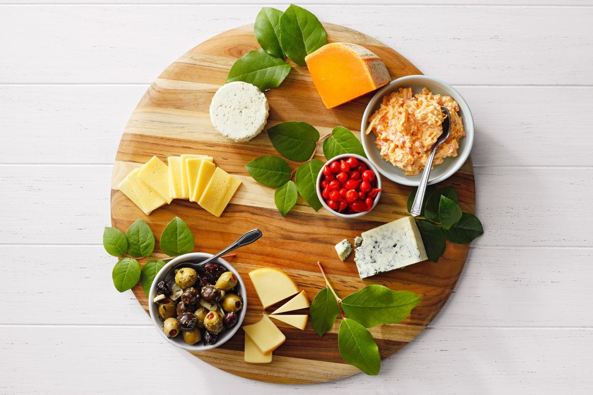 A round wooden board with assorted cheeses, olives, and cherry tomatoes. There's a bowl of cheese spread with a spoon, surrounded by green leaves. The board is on a white wooden surface.