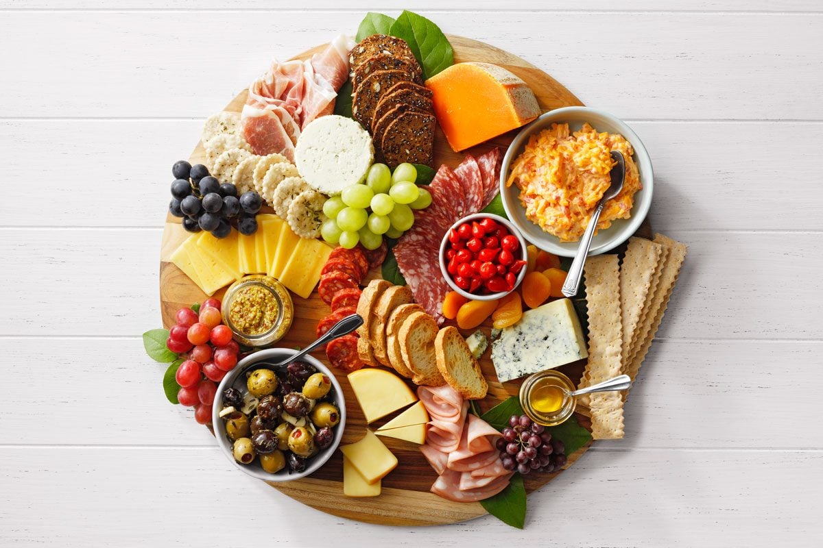 A round wooden platter filled with an assortment of cheeses, meats, crackers, grapes, olives, cherry tomatoes, and dips. The colorful arrangement includes cheddar, brie, salami, and prosciutto, creating a vibrant and appetizing display.