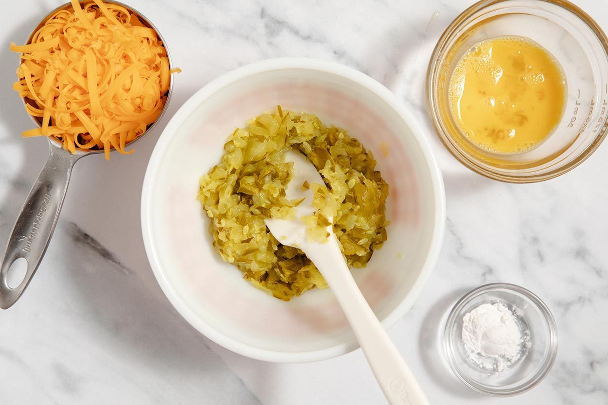 Bowl of chopped pickles, measuring cup of cheddar cheese, small glass bowl of whisked egg, small glass bowl of flour
