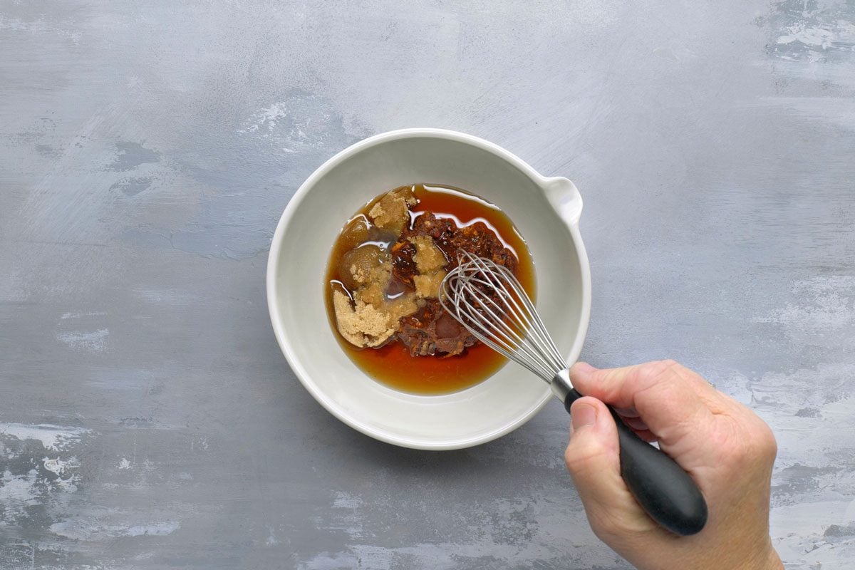 overhead shot of a hand holding a whisk in a white bowl filled with a sauce mixture; The sauce is a light brown color and contains brown sugar and chili flakes; The bowl is sitting on a grey textured surface