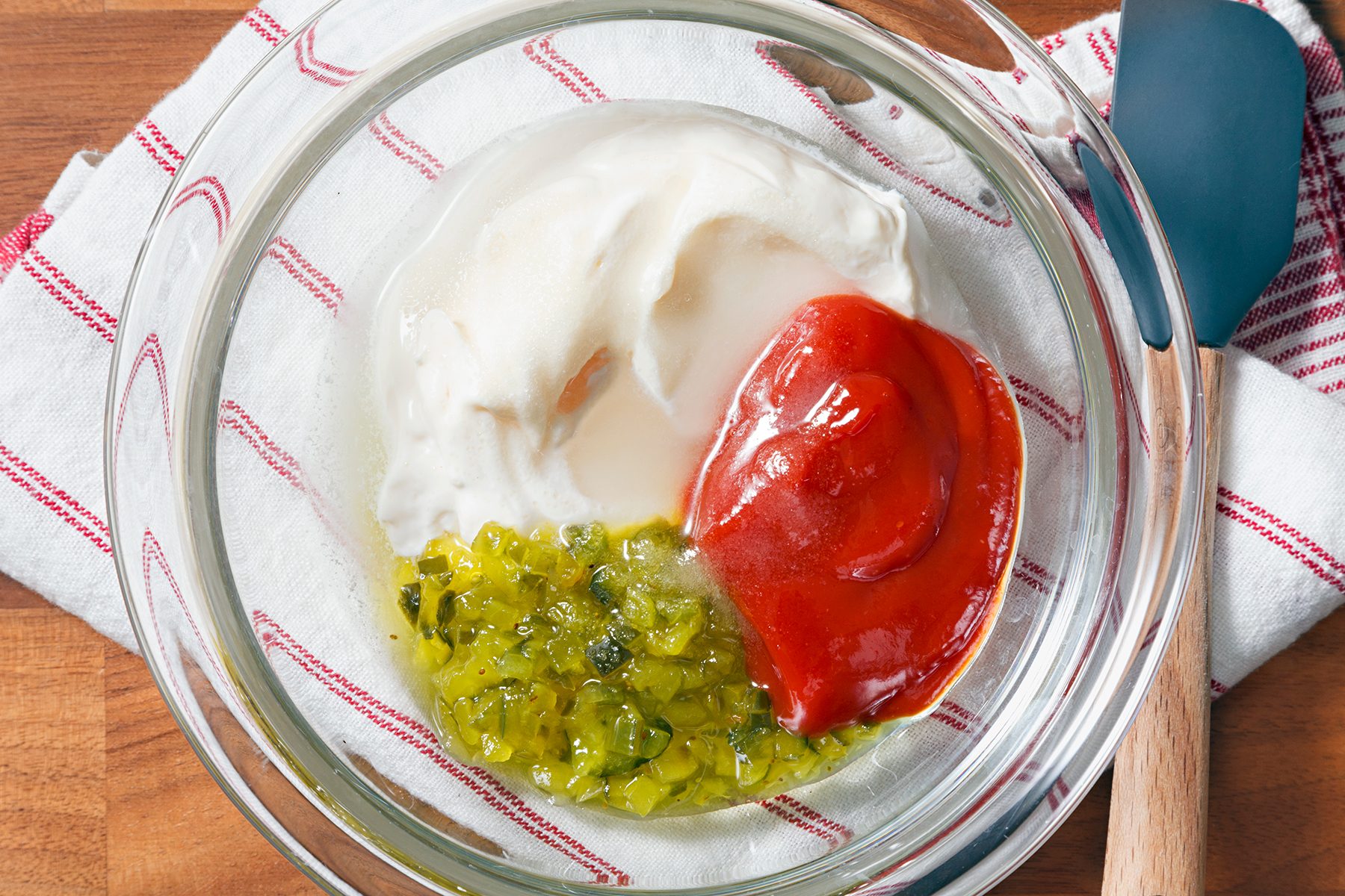A glass bowl containing mayonnaise, ketchup, and chopped pickles on a red and white striped cloth, with a blue spatula and wooden spoon nearby.