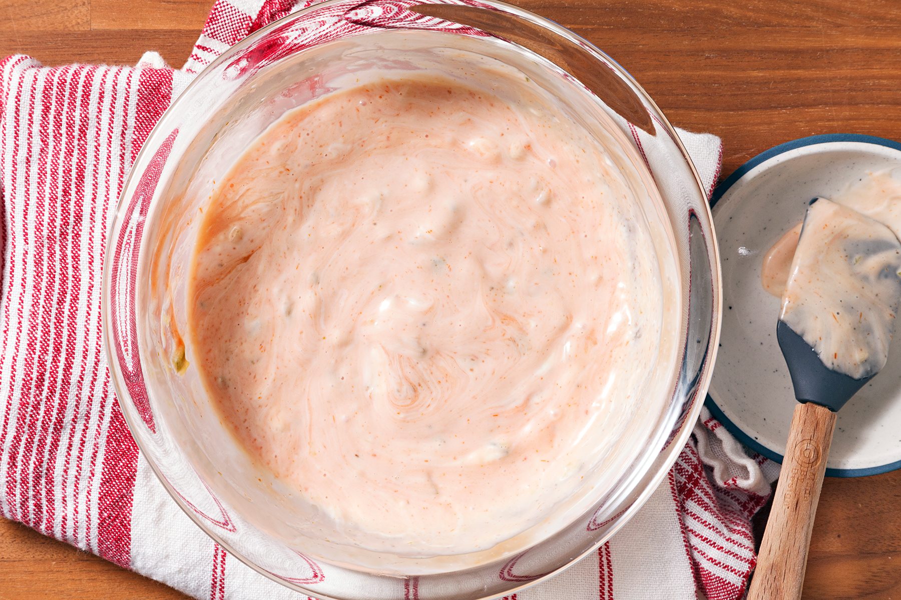 A bowl of dressing in a glass bowl on a red and white striped towel. A spatula with dressing rests on the side of the bowl. The creamy dressing has a light pink hue with visible herbs.