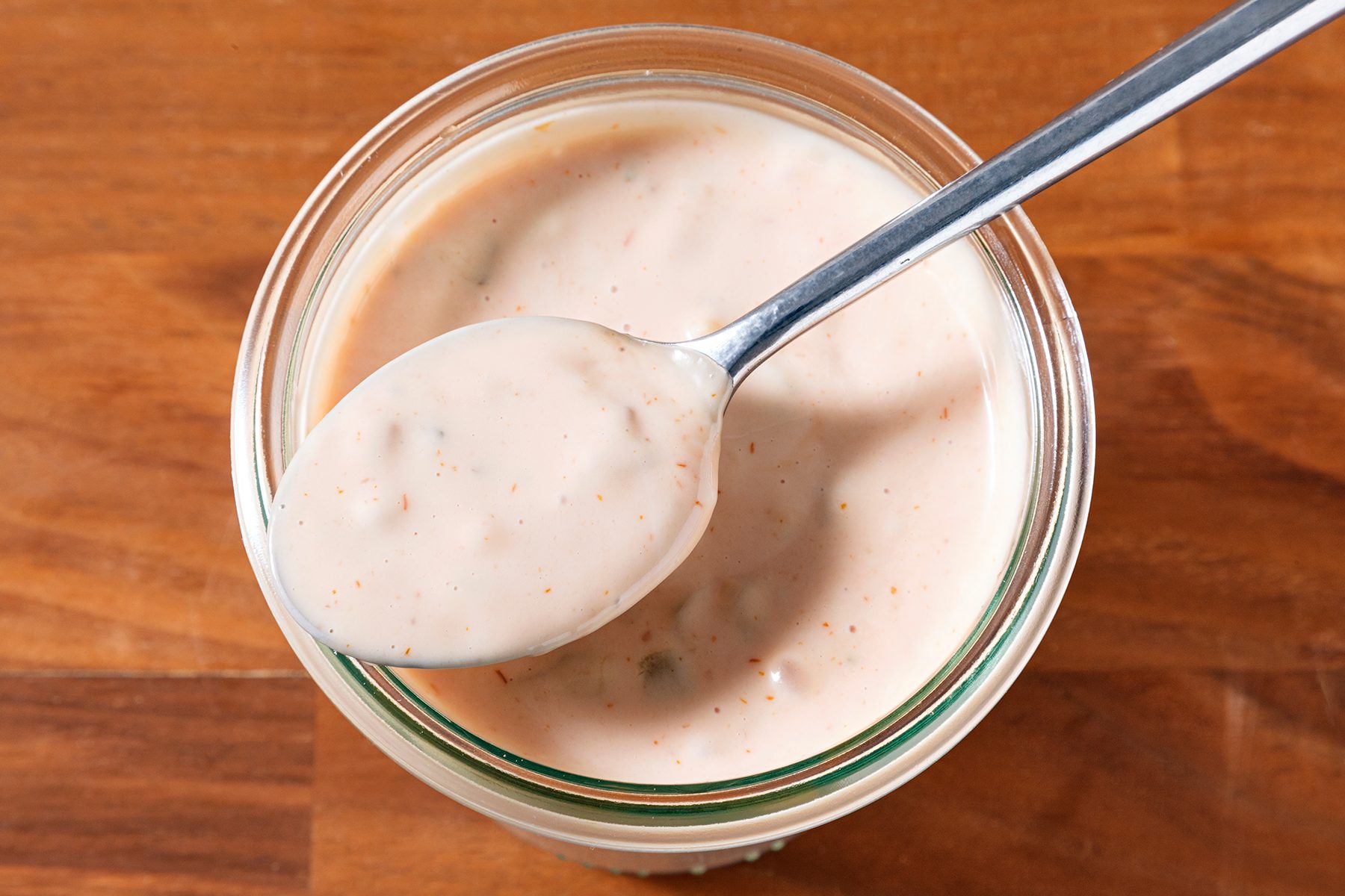A jar of creamy sauce with herbs is shown on a wooden surface. A spoon rests on the jar, displaying the smooth texture of the sauce.