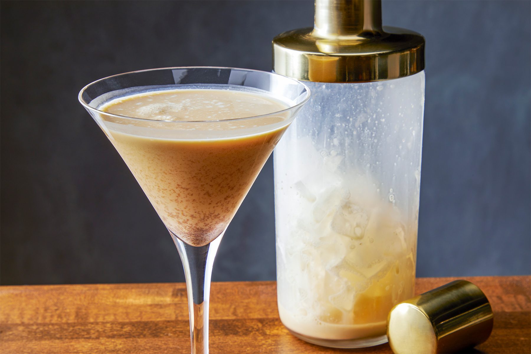 A frothy espresso martini in a martini glass sits on a wooden surface, next to a gold-capped cocktail shaker. The background is dark and blurred, highlighting the drink and shaker.