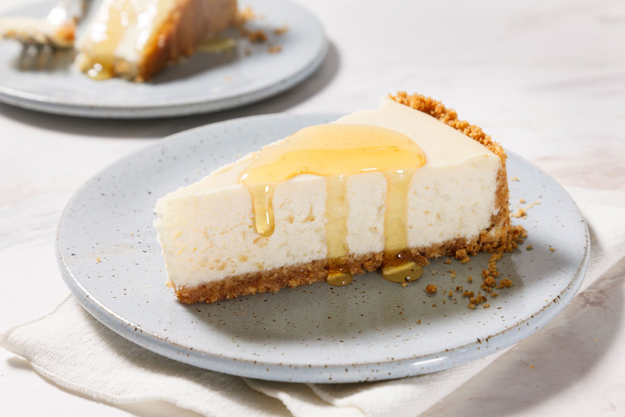 A slice of Goat Cheese Cheesecake with a crumbly base sits on a speckled gray plate. The cheesecake is topped with a drizzle of golden honey. Another piece is blurred in the background. The plate rests on a light fabric napkin.