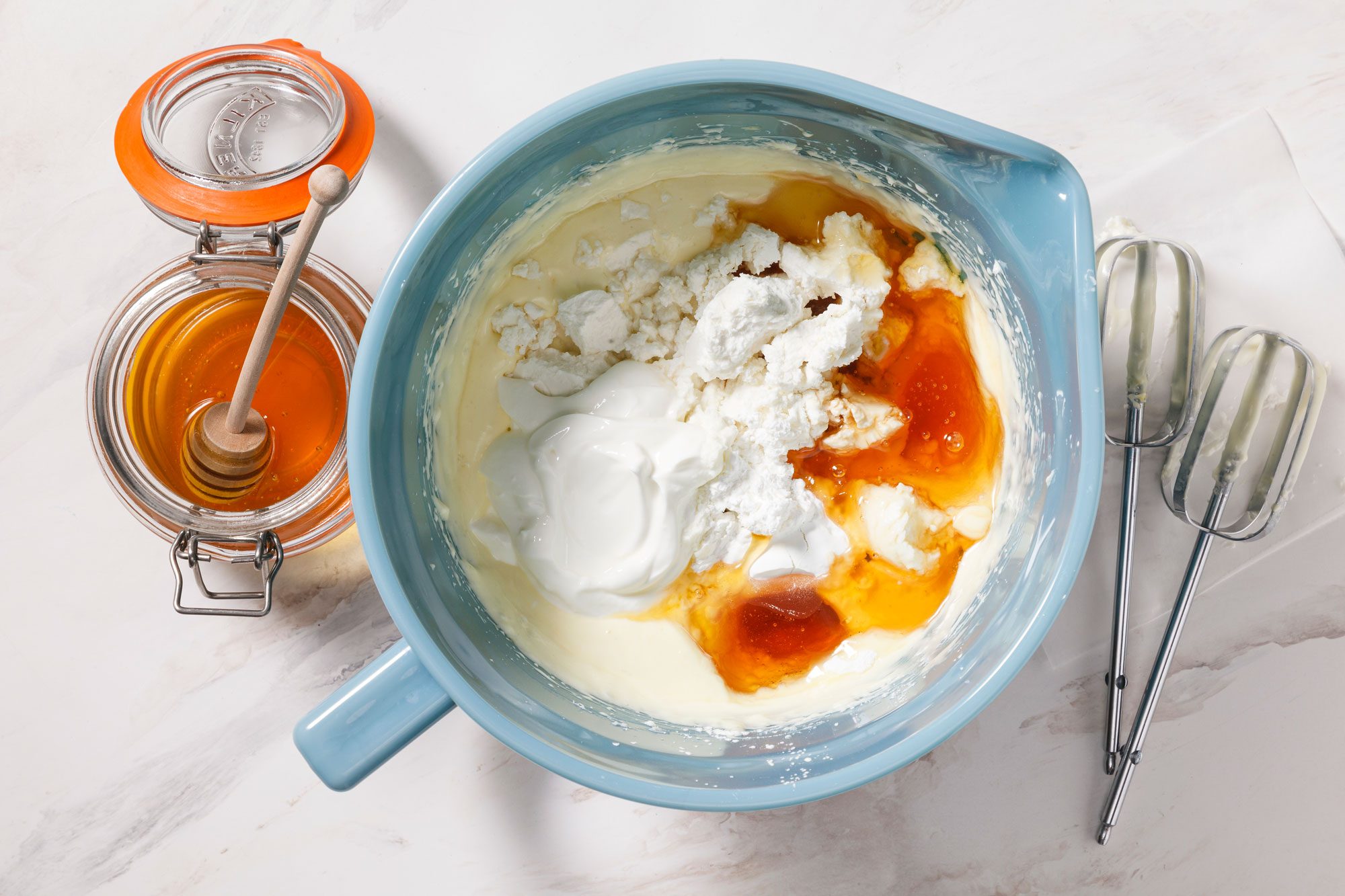 A blue mixing bowl with ingredients, including cottage cheese, yogurt, and honey, on a marble countertop. A jar of honey with a dipper and a pair of electric mixer beaters are next to it.