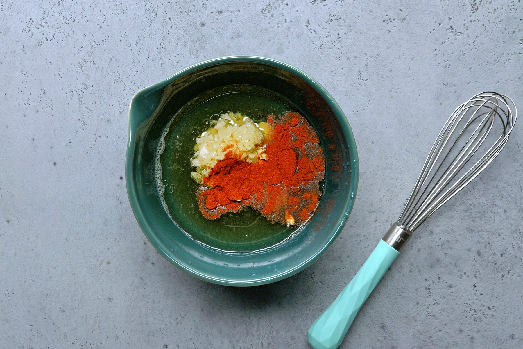 A teal mixing bowl contains chopped garlic, ginger, and paprika powder on a grey countertop. A whisk with a teal handle is placed beside the bowl.