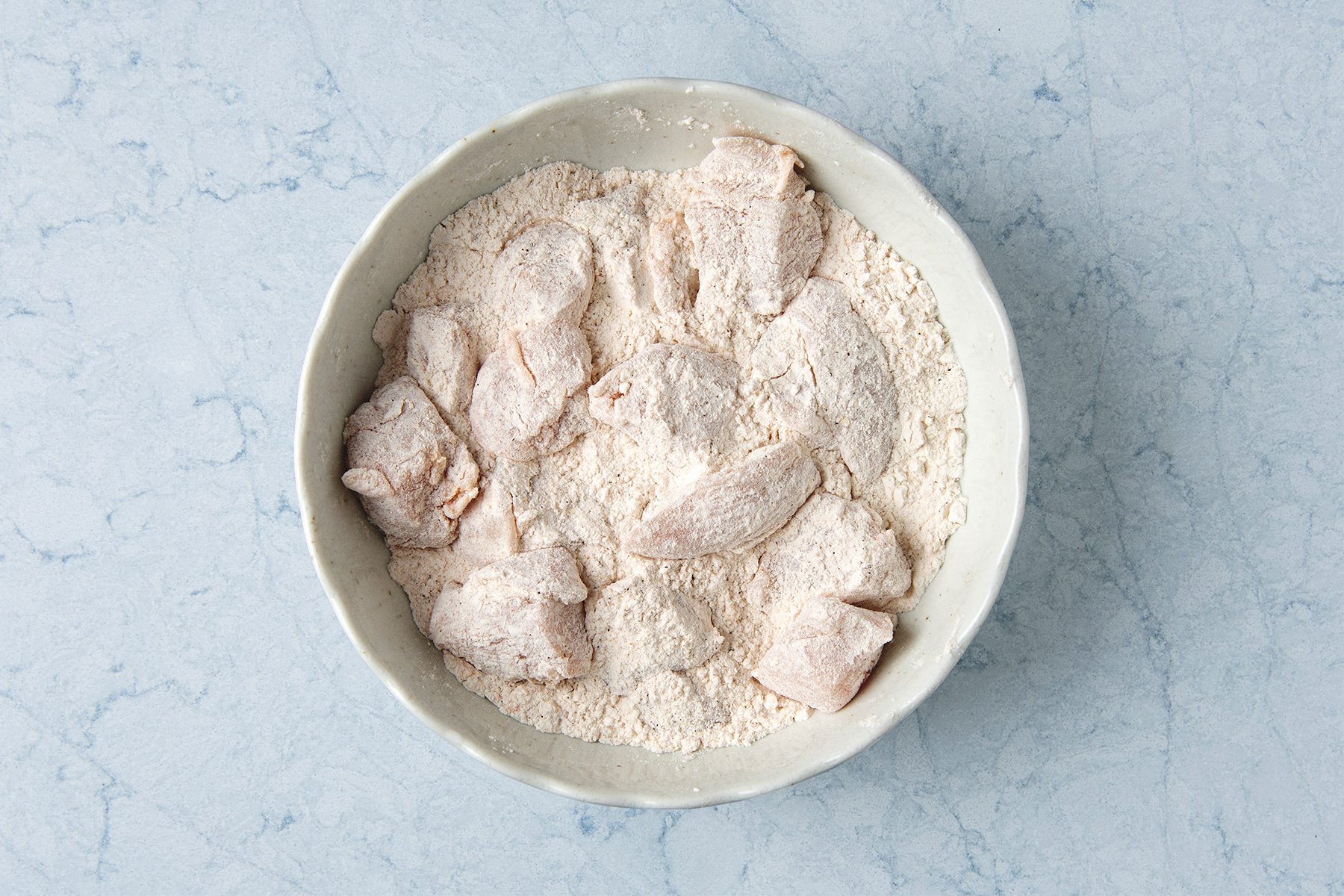 Chicken pieces coated in flour are placed in a white bowl on a light blue marble surface.