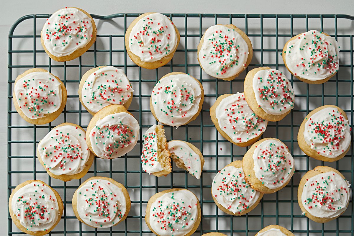 Round sugar cookies with white icing and red and green sprinkles are arranged on a cooling rack. One cookie is broken in half, revealing a soft interior. The rack is placed on a white surface.