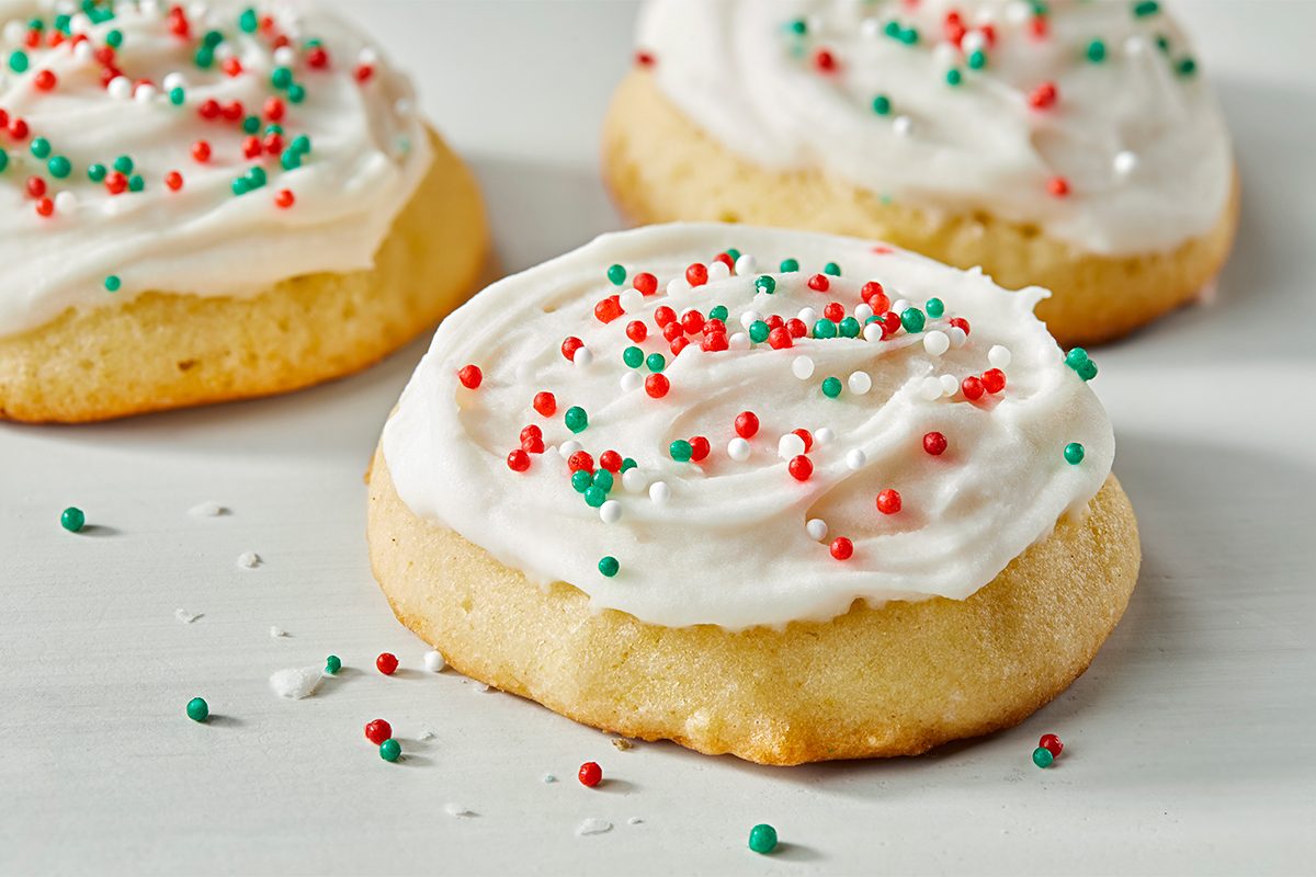 Three sugar cookies topped with white frosting and red, green, and white sprinkles are arranged on a light surface.