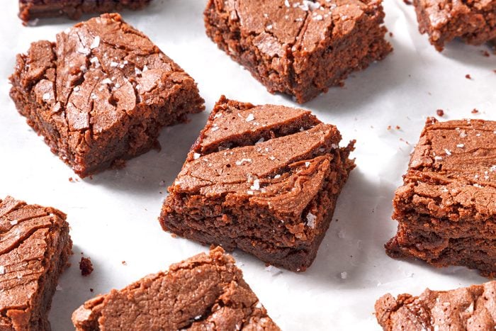 Squares of chocolate brownies sprinkled with sea salt are arranged on a white parchment paper. The brownies have a rich, fudgy texture with slightly cracked tops.