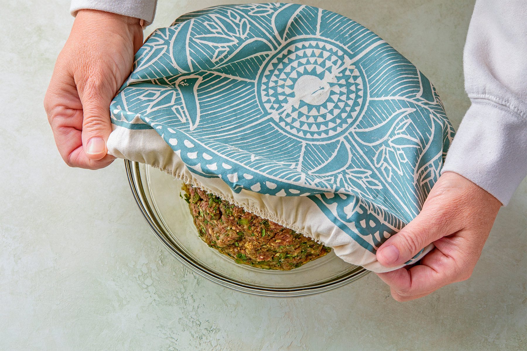 overhead shot of a person's hands are holding a fabric over a bowl of meatloaf mixture; the fabric is a light blue and white with a geometric pattern; the bowl is glass and the meatloaf mixture is made of ground meat and herbs