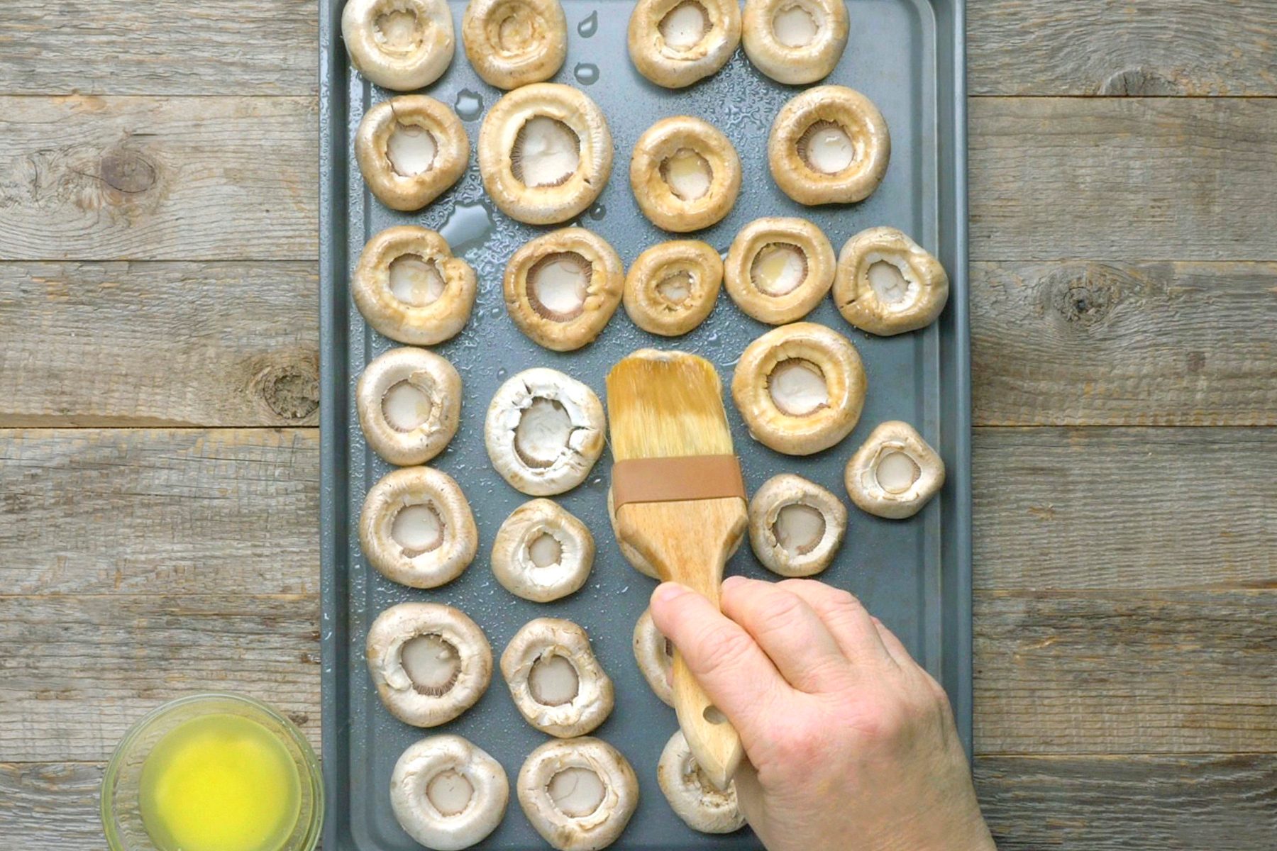 A hand brushes mushroom caps with oil on a baking sheet set on a wooden surface.