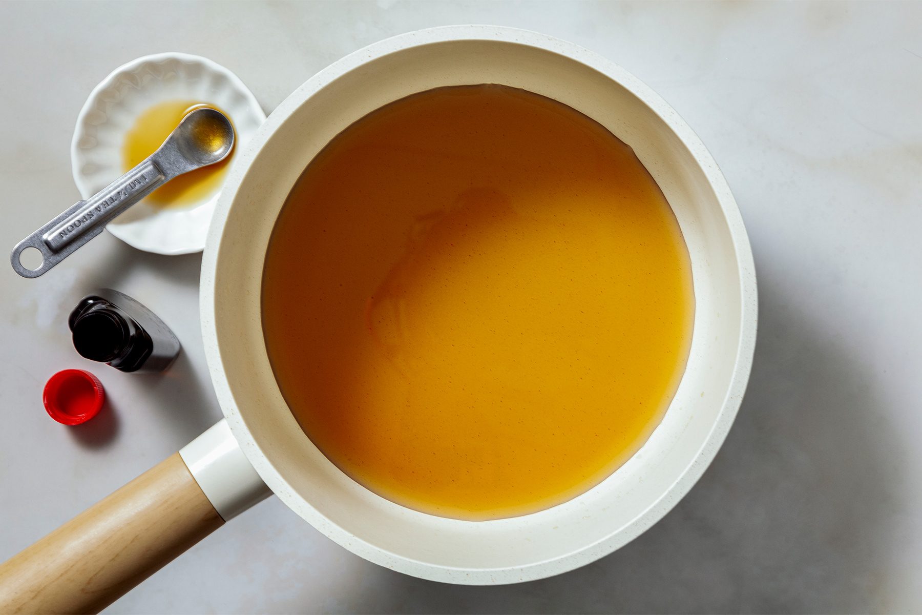 A saucepan filled with a smooth orange sauce on a light surface. Nearby, there are two small bottles, one with a red cap, and a teaspoon on a small white dish.
