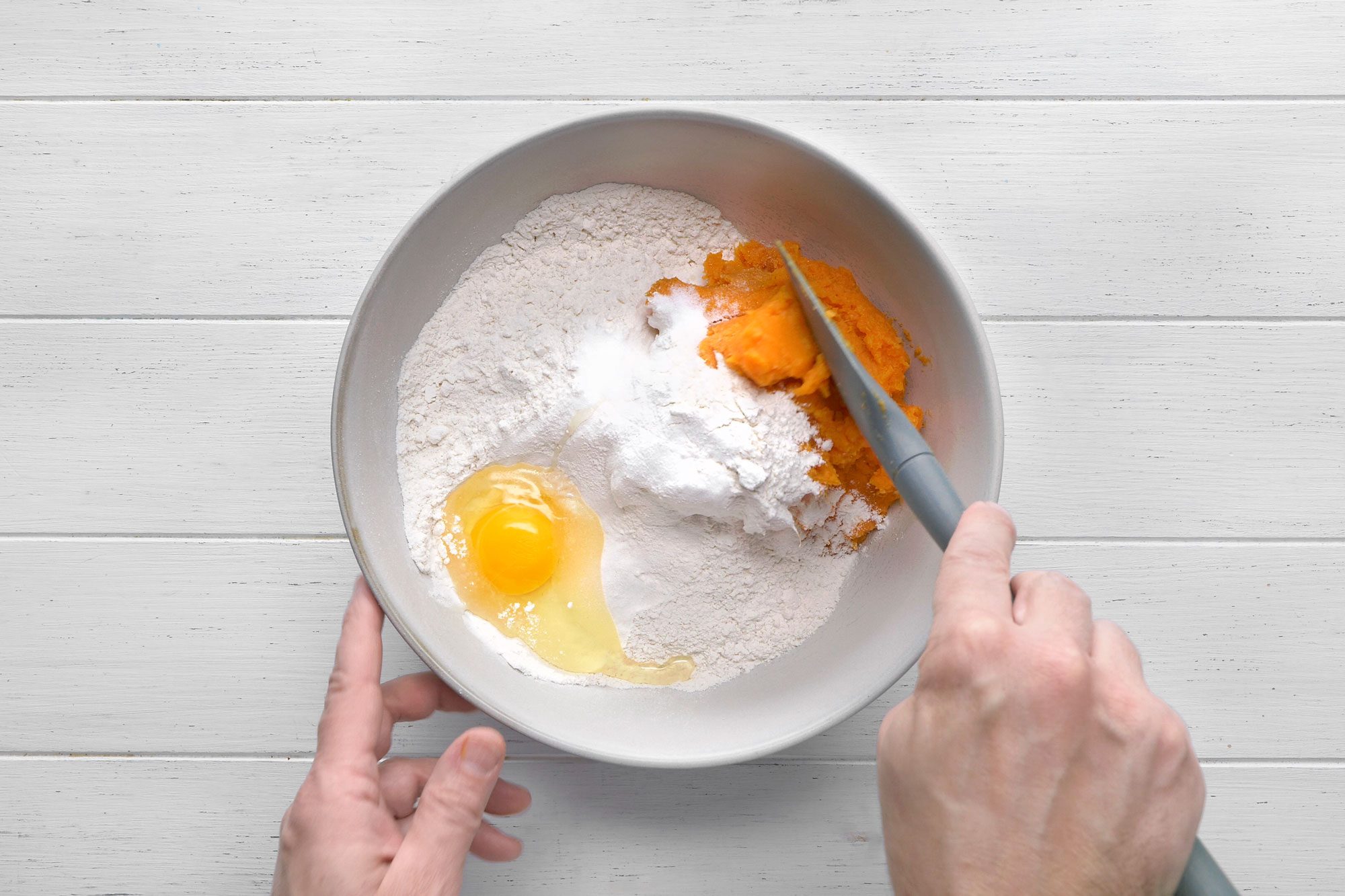 overhead shot of a white bowl with ingredients for baking; the ingredients in the bowl are flour, an egg, sugar, and mashed sweet potatoes; the bowl is sitting on a white wood table