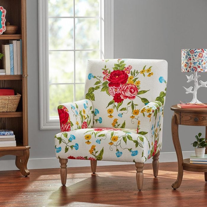White armchair with colorful floral designs sits on a wooden floor with a gray wall, window, side table, and bookshelf in the background
