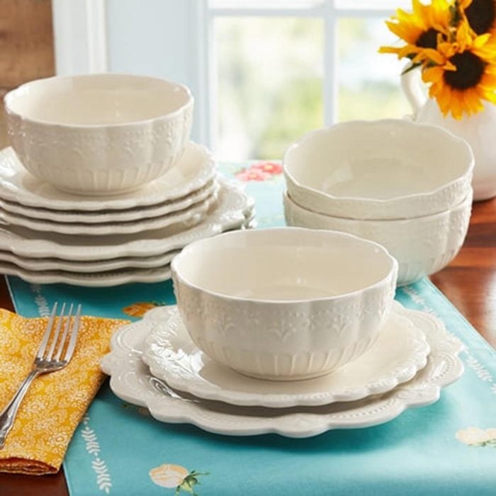 Several stacked, white, scalloped dinner plates and bowls on a teal place mat with a yellow napkin and silver fork, with a window and vase of sunflowers in the background