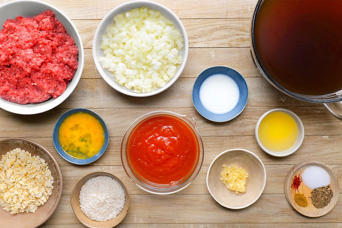 Various cooking ingredients on a wooden surface: ground beef, chopped onions, beef broth, salt, melted butter, tomato sauce, egg mixture, minced garlic, breadcrumbs, flour, and spices in separate bowls and dishes.