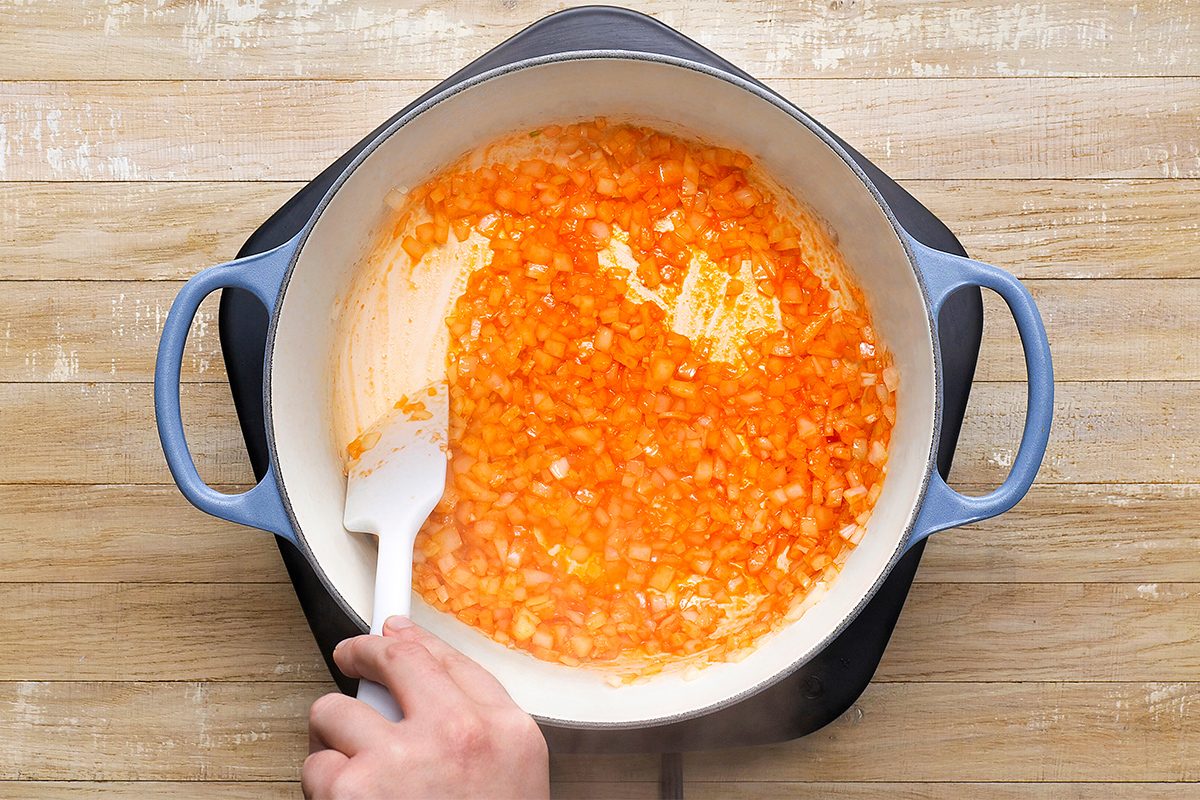 A person stirs chopped onions sautéing in a large blue pot on a stovetop. The onions appear to be partially caramelized, giving a golden orange hue. The pot is set on a wooden surface.