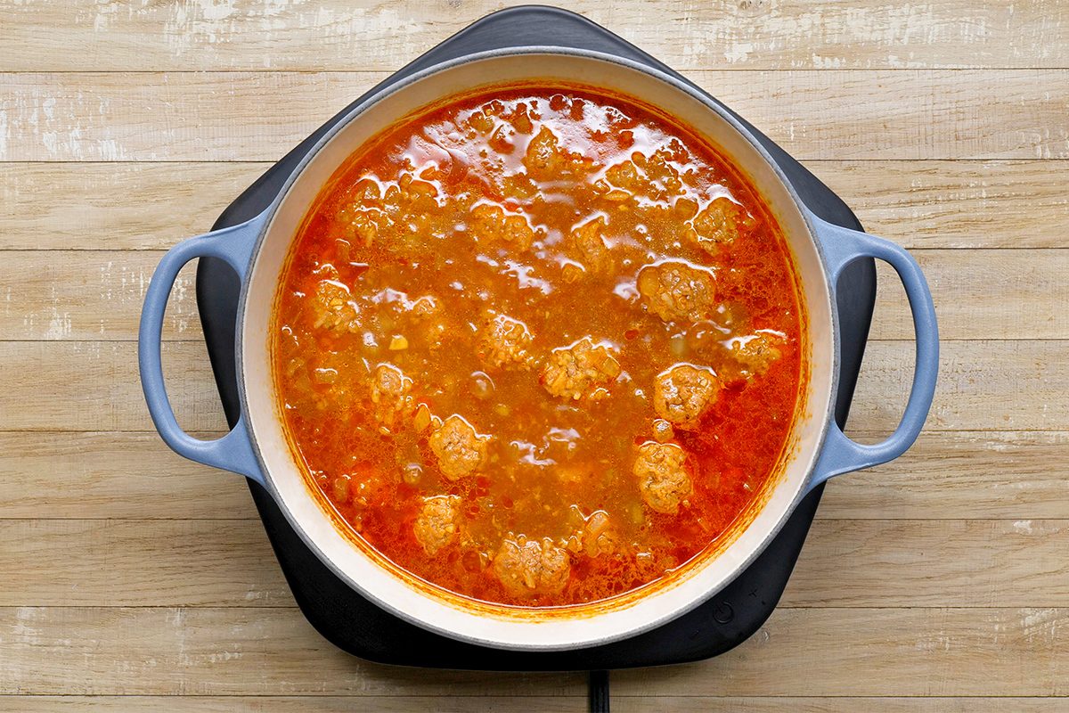 A pot filled with a thick, red-orange stew containing visible chunks of meat is placed on a wooden surface. The pot has two handles and is resting on a trivet.
