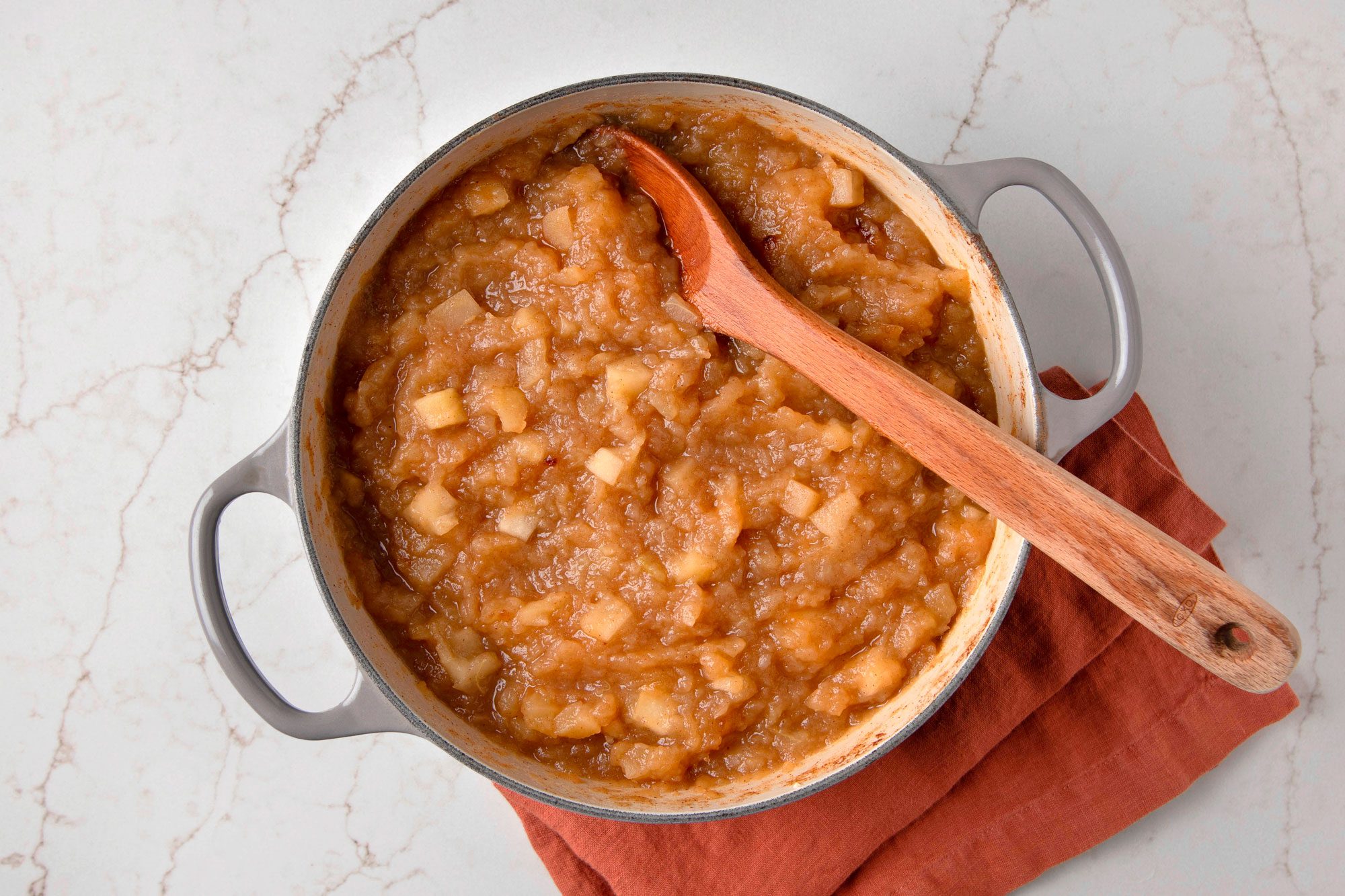 Overhead shot of a pot of applesauce, a wooden spoon, and a maroon kitchen towel, all on a white marble counter