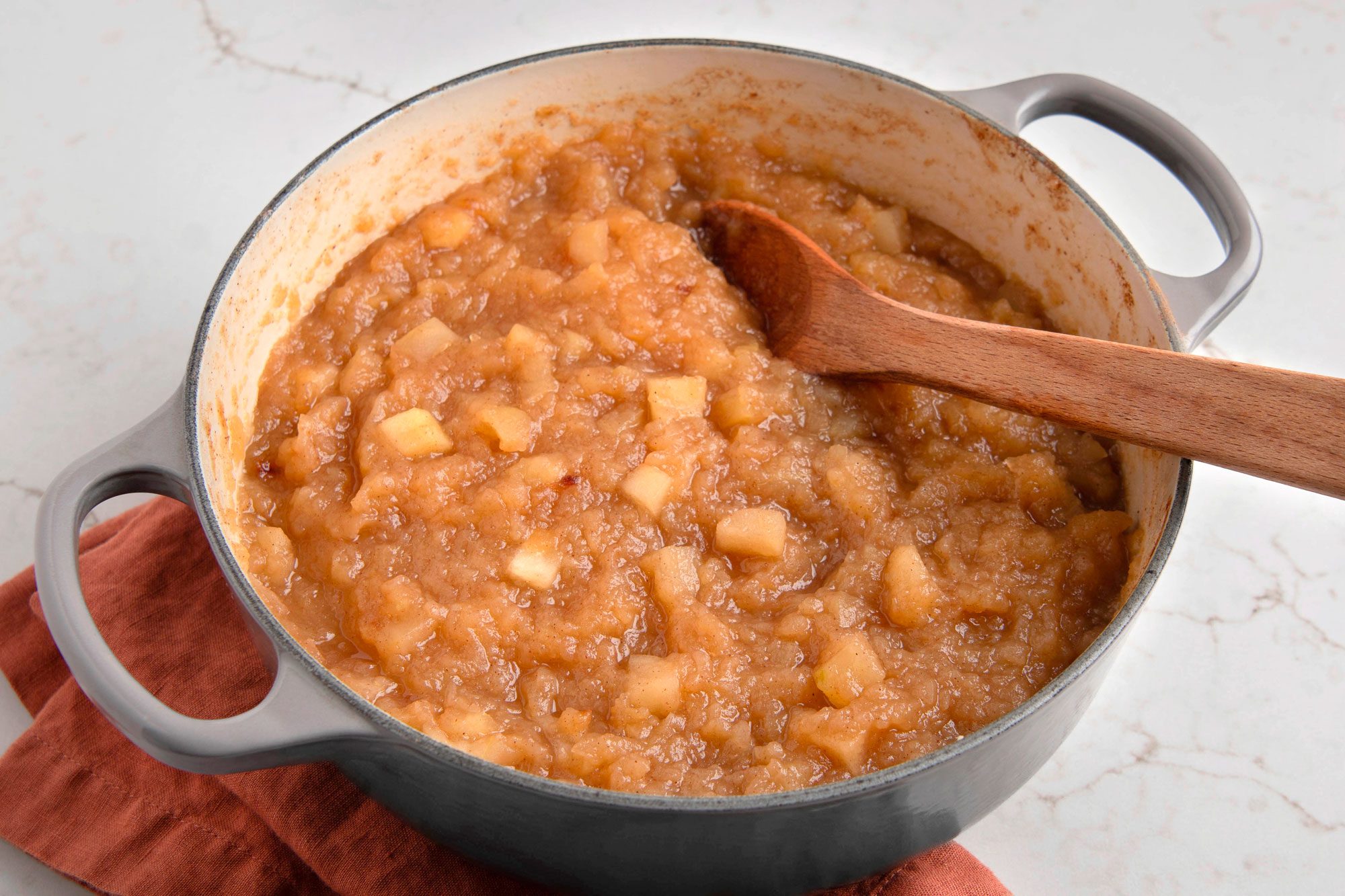 The pot is gray with a brown handle, the applesauce is a light brown color and has some visible pieces of apple in it; the applesauce is sitting on a brown napkin and a white marble counter