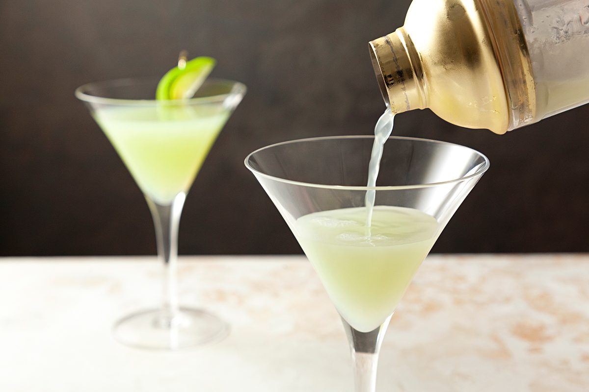 A golden shaker pours a pale green cocktail into a martini glass. Another martini glass with a similar drink and an apple slice garnish is in the background on a light-colored surface.
