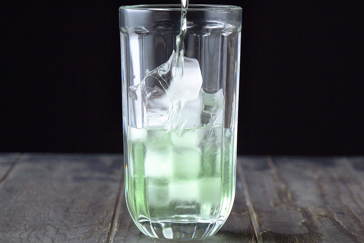 A clear glass filled with ice cubes is being poured with a light green liquid. The glass is set on a dark wooden surface against a black background.