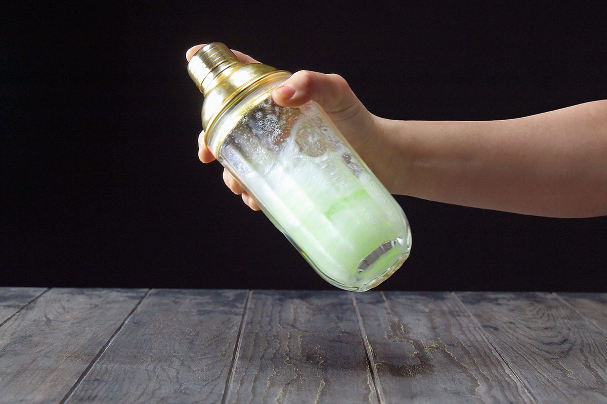 A hand holds a cocktail shaker with a golden top, vigorously shaking it. The shaker is filled with a light green liquid. The background is dark, and the surface is a textured wooden table.