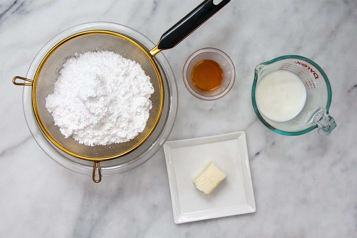 Ingredients arranged on a marble surface: sifted powdered sugar in a sieve, a small bowl of vanilla extract, a measuring cup with milk, and a square plate with a pat of butter.