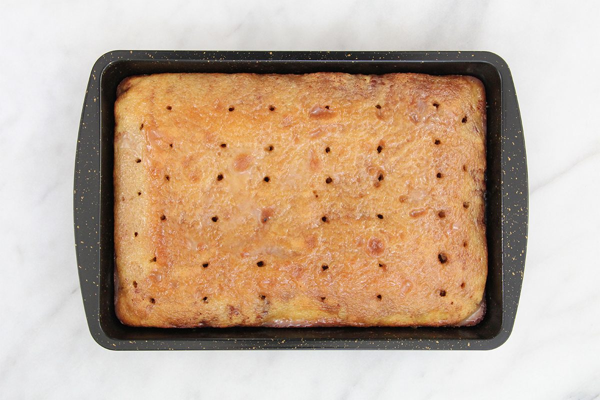 A rectangular cake with holes on top sits in a black baking tray. The cake appears lightly browned and evenly baked, placed on a white marble surface.