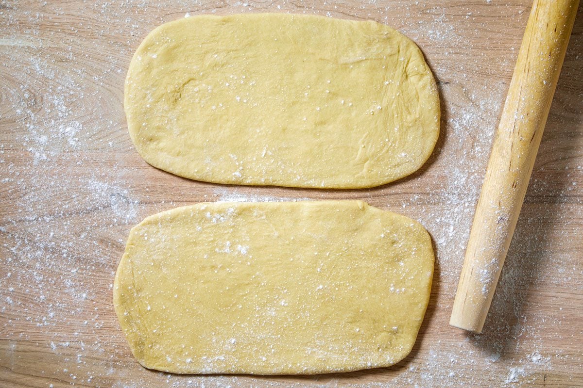 overhead shot of a wooden cutting board, dusted with flour, with two pieces of dough and a rolling pin;
