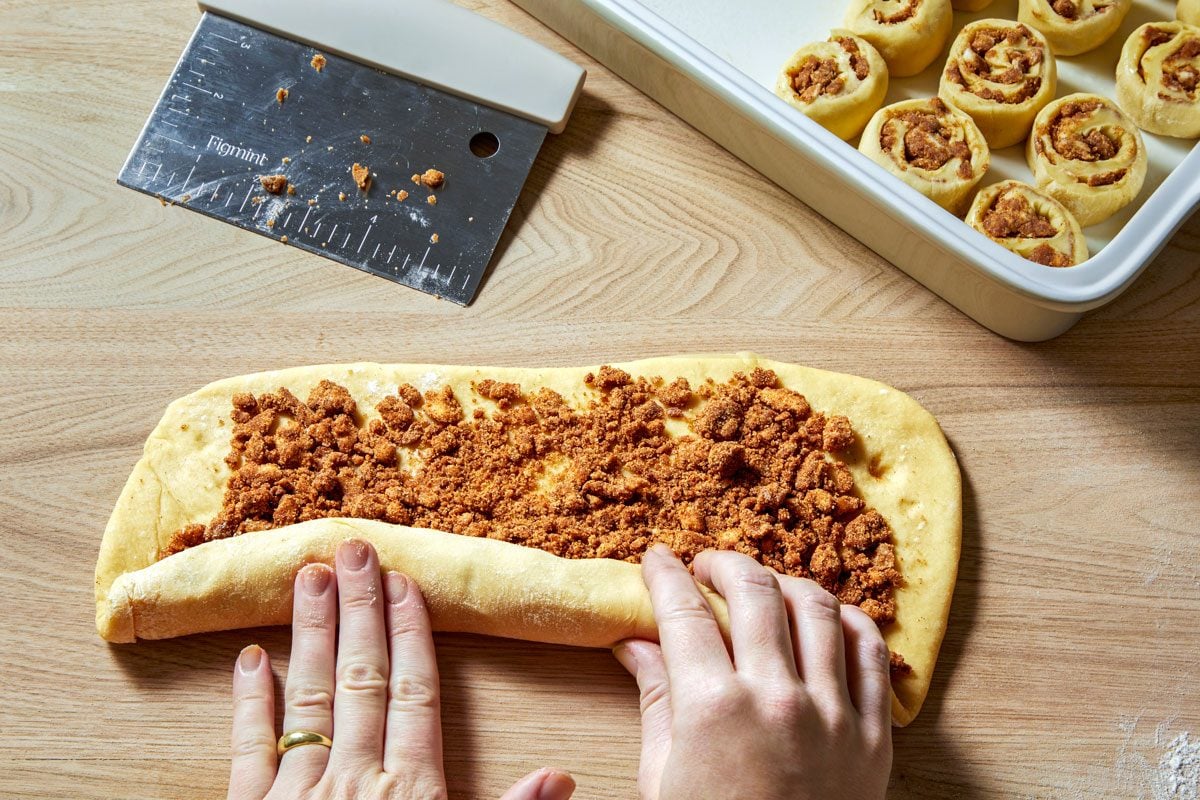 3/4th shot of a person rolling up a piece of dough that is covered in cinnamon sugar; the dough is being rolled up on a wooden surface; a white baking dish filled with already rolled up cinnamon rolls is shown in the upper right-hand corner of the image; a metal pastry scraper with a white handle is shown in the upper left-hand corner of the image