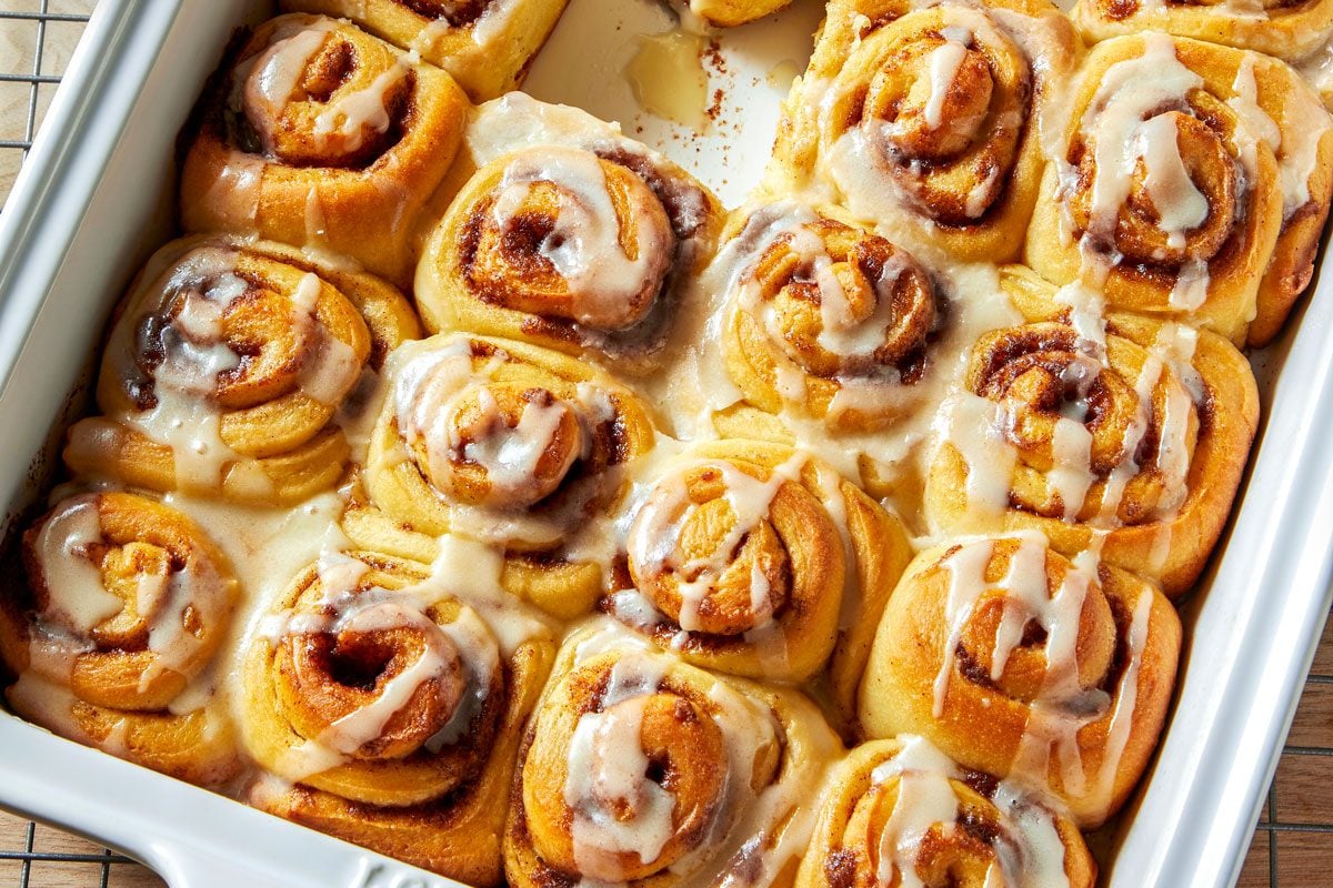overhead shot of a white baking dish filled with freshly baked cinnamon rolls, topped with a creamy white icing; the dish is sitting on a wire rack, and a small bowl of icing is on the left side of the frame;