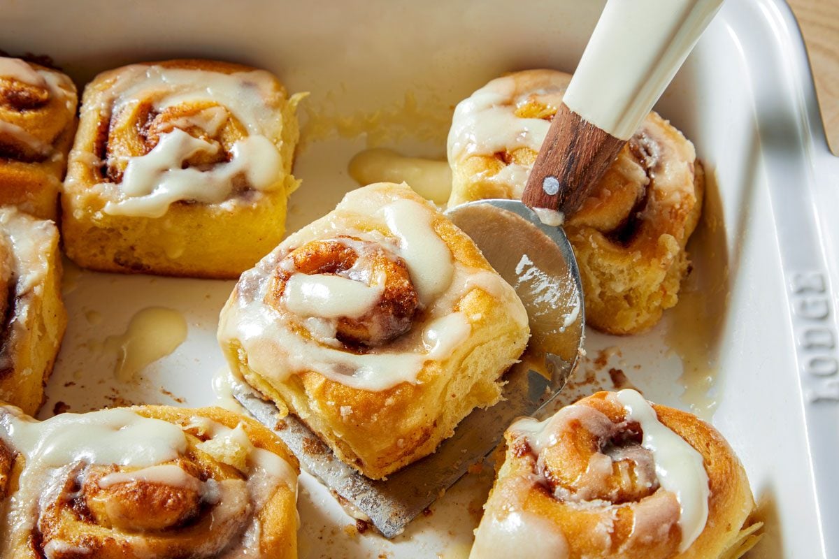 a white baking dish filled with freshly baked cinnamon rolls; the rolls are covered in a thick layer of white frosting and there is a pool of melted frosting at the bottom of the baking dish; a silver metal spatula with a wooden handle is used to cut the cinnamon rolls, and a piece of cinnamon roll is being lifted off the baking dish by the spatula