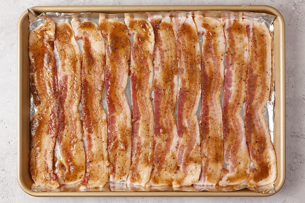 Strips of bacon are arranged in rows on a baking sheet lined with foil, seasoned with brown sugar and spices, ready for cooking.