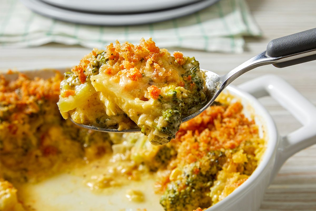 Side view of Cheesy Broccoli Au Gratin kept in a baking dish on a kitchen cloth with a serving being taken out with a metal spoon.