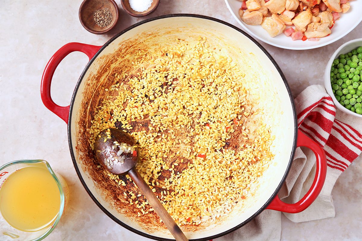 A red pot on a light surface contains sizzling garlic and spices. Nearby, a wooden spoon rests in the pot. Surrounding the pot are bowls with diced chicken, peas, seasoning, and a measuring cup of broth. A striped kitchen towel is also visible.