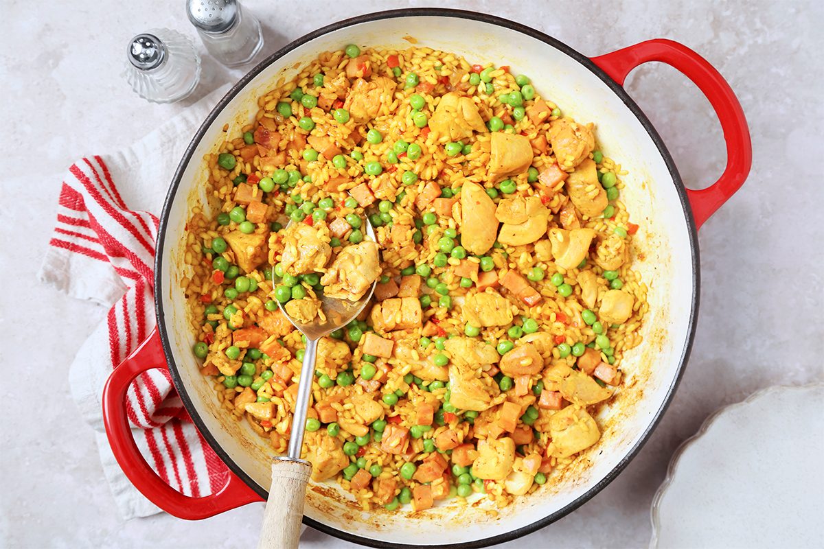 A large red pan filled with a vibrant dish of chicken fried rice, featuring diced chicken, peas, and carrots. A spoon rests inside. A red-and-white striped cloth and salt and pepper shakers are nearby on a light surface.