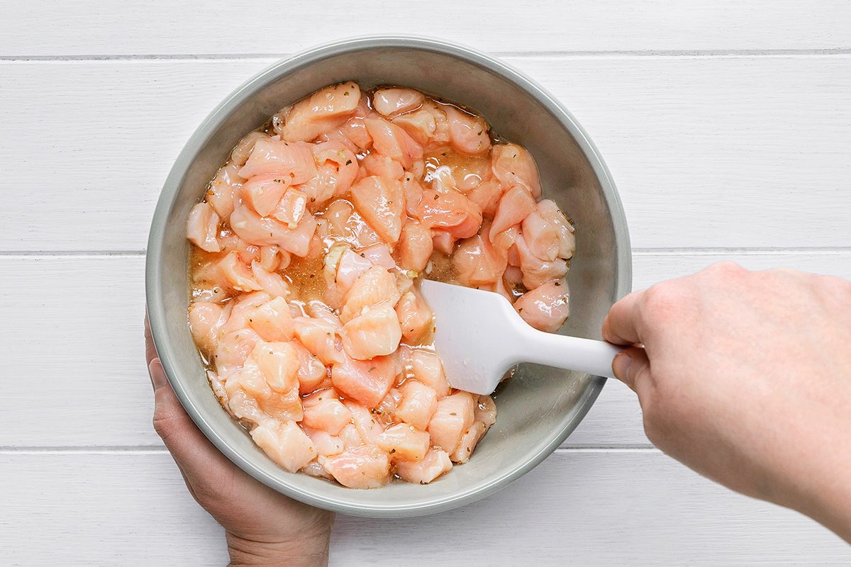 Person stirring raw, diced chicken in a bowl with a white spatula on a white wooden surface.