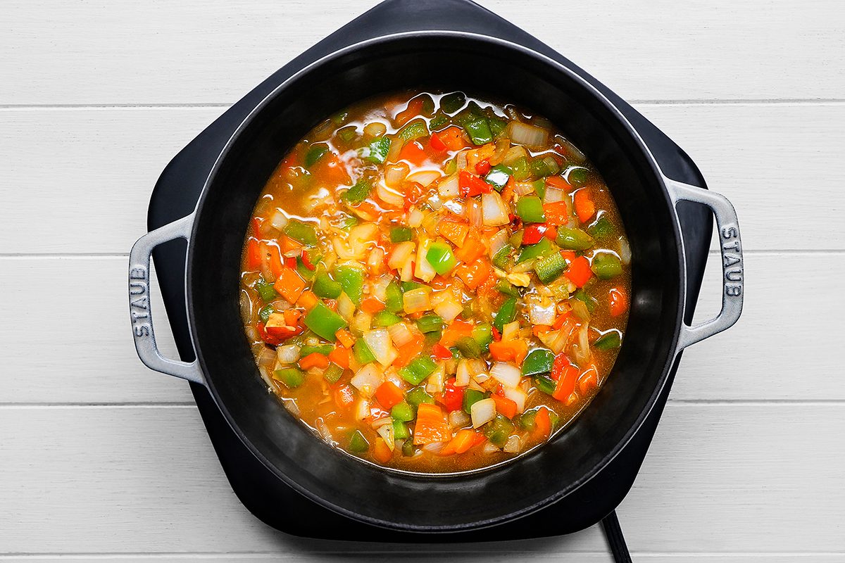 A black Staub pot filled with a colorful vegetable soup, featuring chopped carrots, bell peppers, onions, and a simmering broth. The pot sits on a white wooden surface.