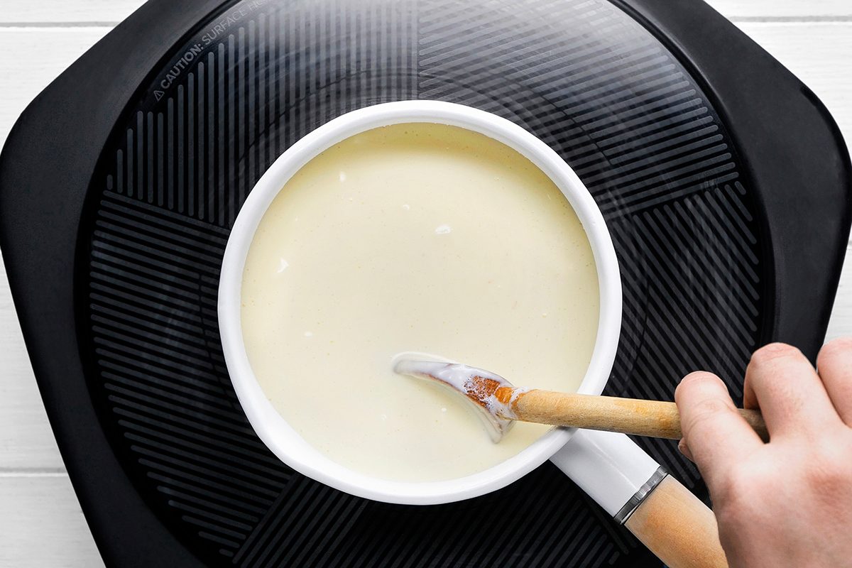 A person's hand stirring a creamy white sauce in a white saucepan with a wooden spoon on a black stovetop.