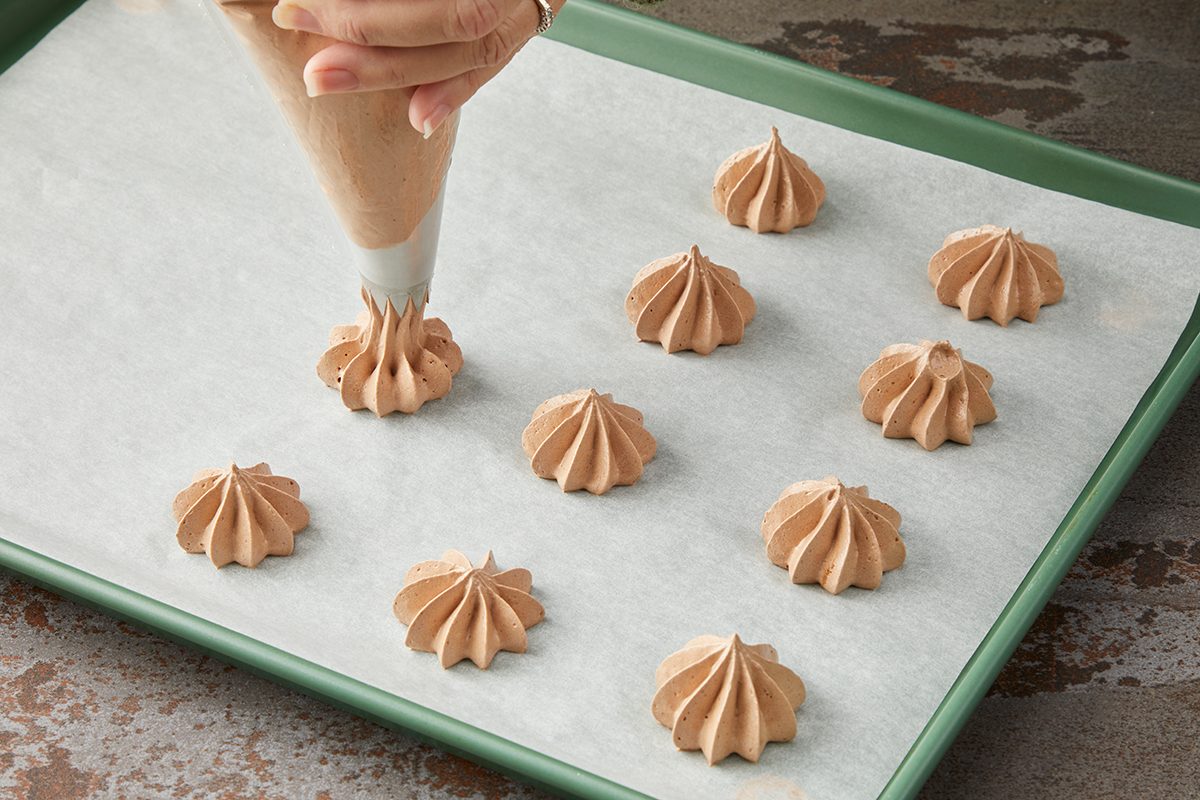 Close-up of a hand using a piping bag to create small, swirled meringue cookies on a parchment-lined baking sheet. The cookies are evenly spaced and have a light brown color.