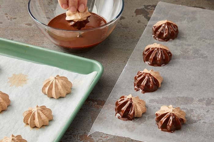 A hand dips meringue cookies into a bowl of melted chocolate. Some meringues are set on wax paper with chocolate coating partially covering them, while others are on a green tray lined with parchment paper.
