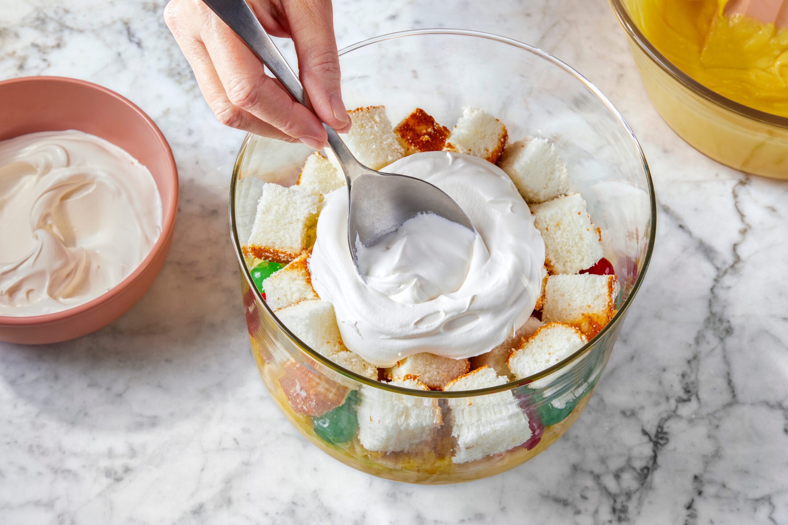 A hand holding a spoon is spreading whipped cream over a layered dessert in a glass bowl. The dessert includes pieces of white cake and colorful gelatin. A bowl with more whipped cream is on the side. The scene is set on a marble countertop.